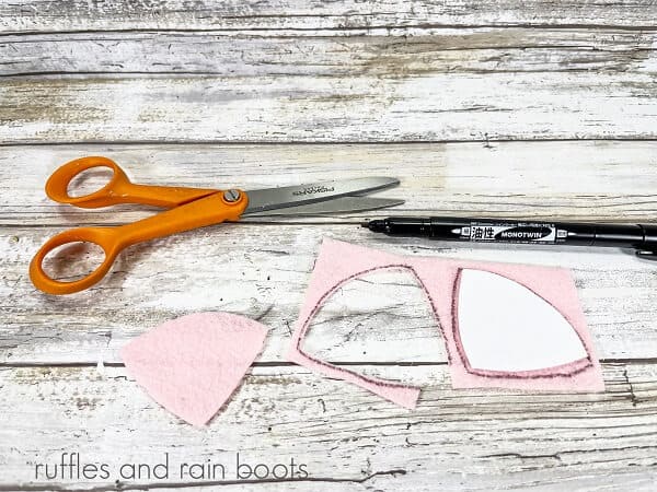 Pink felt next to a black marker and orange handled scissors on a white weathered wood surface.