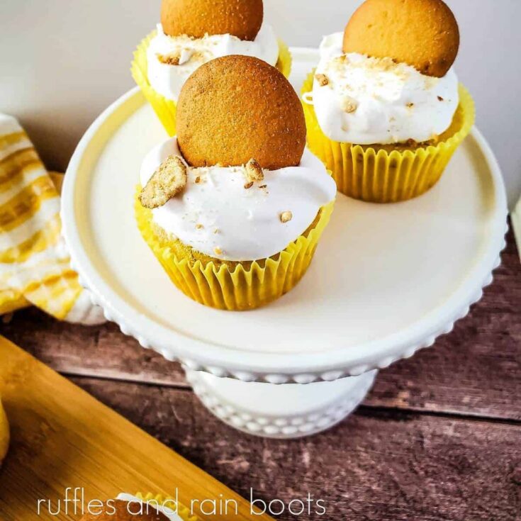 3 banana cream pie cupcakes on a white cake stand next to a wooden cutting board and yellow and white towel on a weathered wood background.
