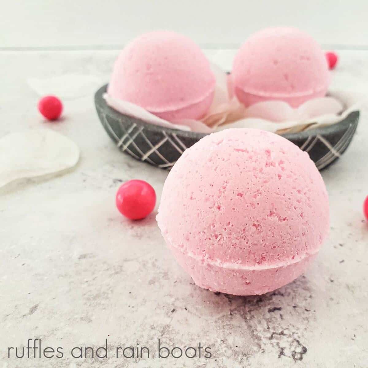 Close-up image of a large pink bath bomb in front of a small black and white bowl with two smaller bath bombs, next to several bubblegum balls and faux flower petals on a white concrete background.