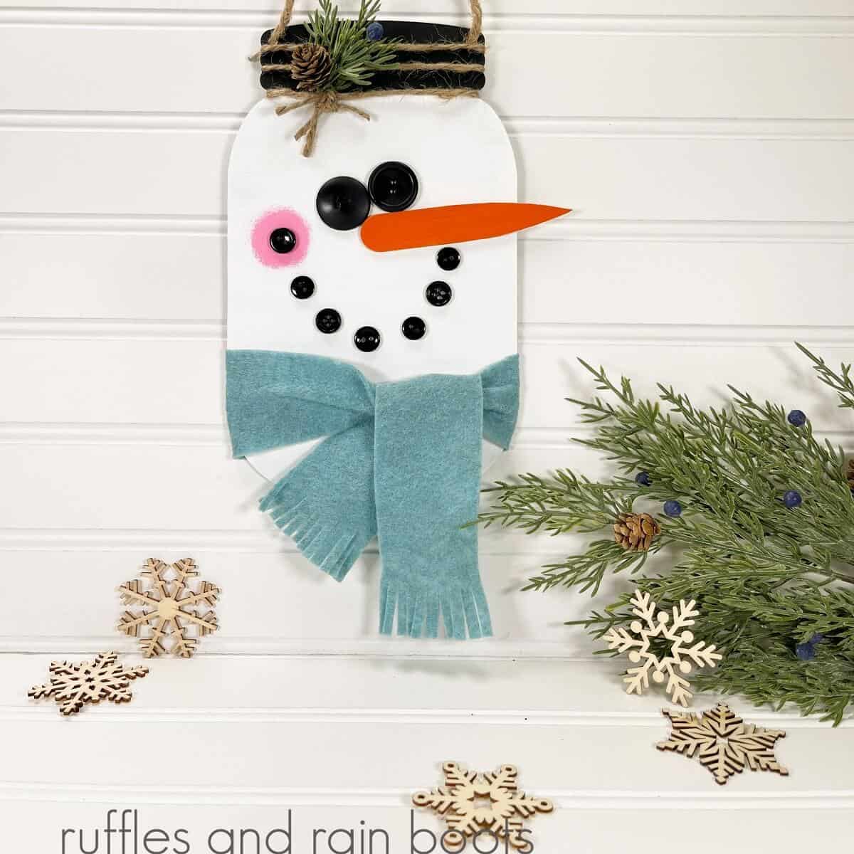 square image of a painted galvanized metal Hanging Mason Jar Snowman next to a piece of faux greenery and 5 wooden snowflakes on a white beadboard background