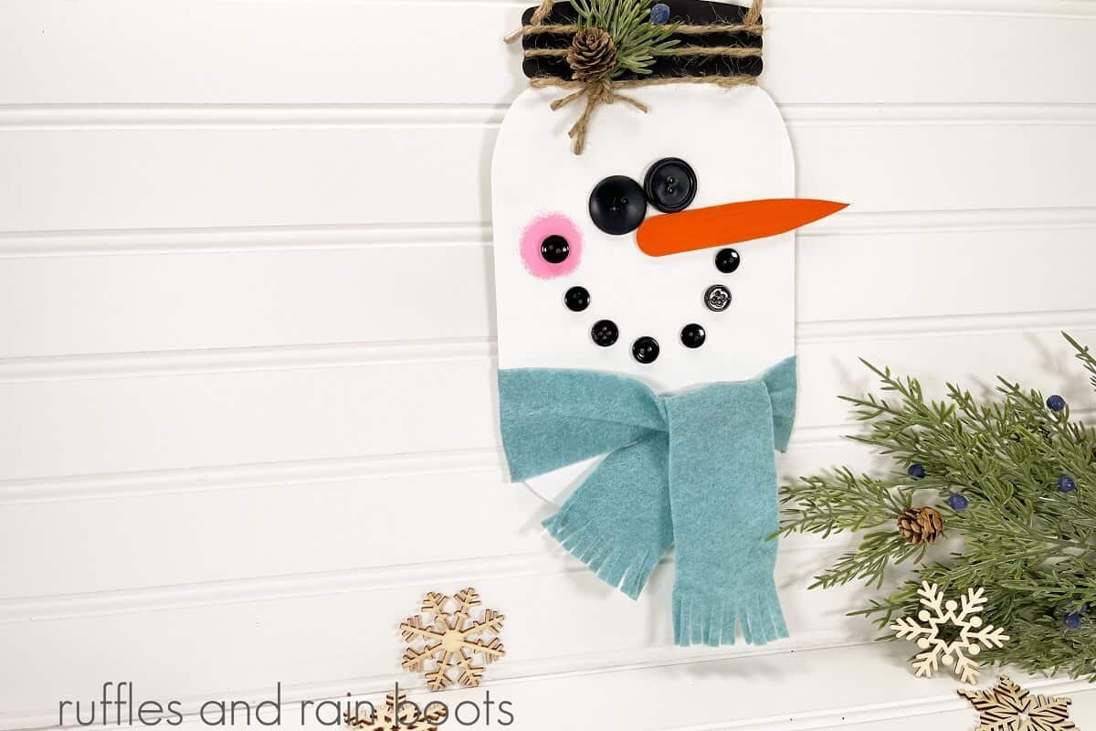 horizontal image of a painted galvanized metal Hanging Mason Jar Snowman next to a piece of faux greenery and wooden snowflakes on a white beadboard background