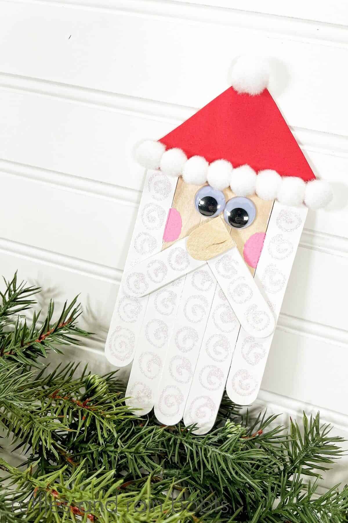 A vertical image of a completed Craft Stick Santa DIY on top of faux holiday greenery and pinecones against a white beadboard wood background.