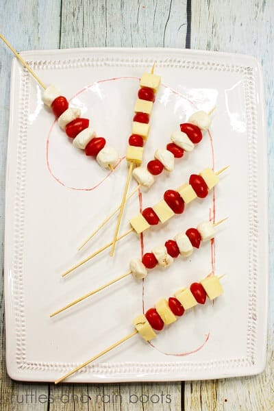 A candy cane shape drawn on a white plate with cheeses on tooth picks against a white weathered wood backdrop.