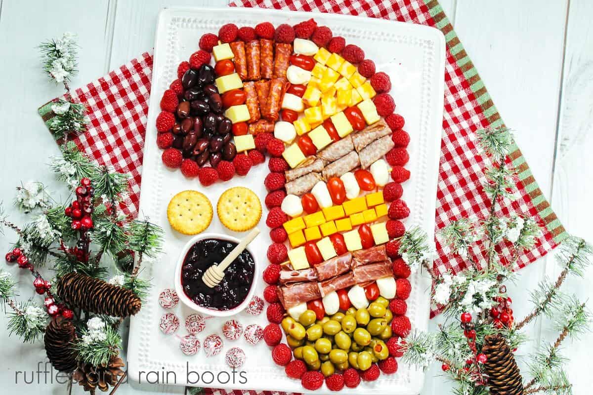 A horizontal image of the meat, cheese, olives, jam and fruit charcuterie board in the shape of a candy cane, on a white plate against a red and white gingham towel on a weathered white wood backdrop, next to faux winter greenery with pine cones.