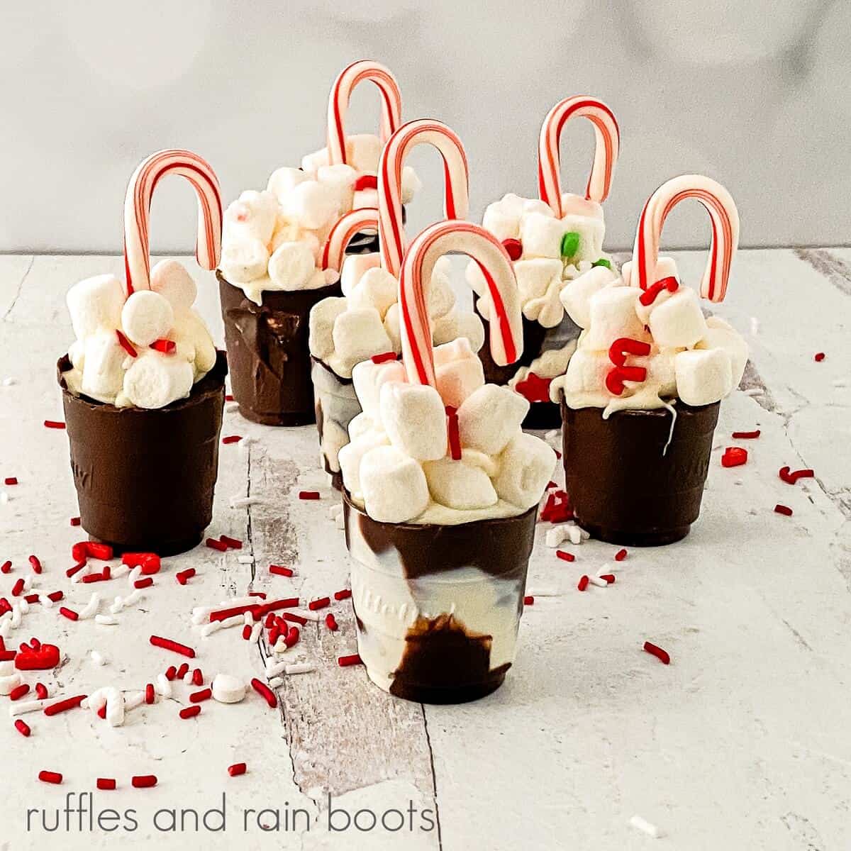 A square image of a group of decorated Hot Cocoa Cup Shots against a white backdrop with holiday sprinkles scattered around them against a white backdrop.