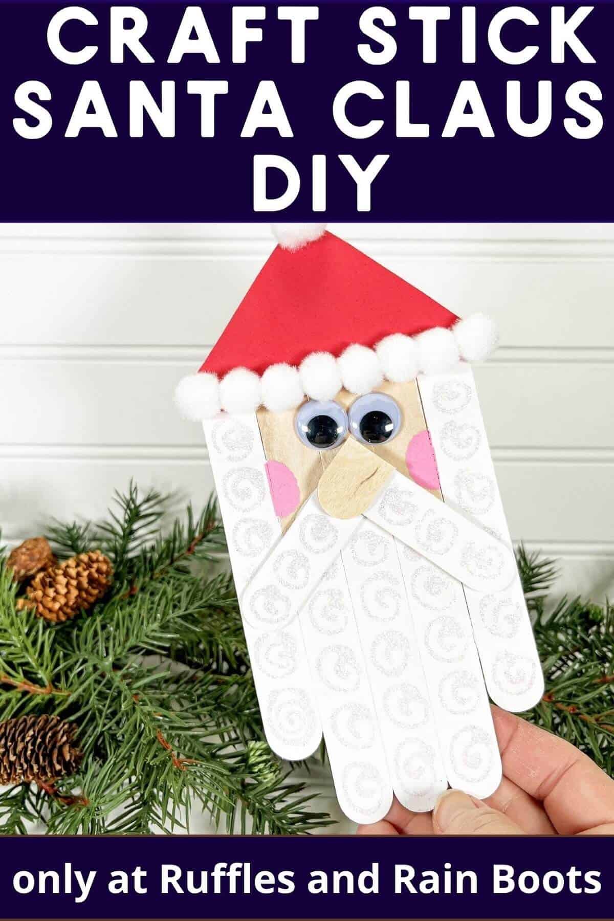 A vertical image with a hand holding a completed Craft Stick Santa DIY on top of faux holiday greenery and pinecones against a white beadboard wood background.