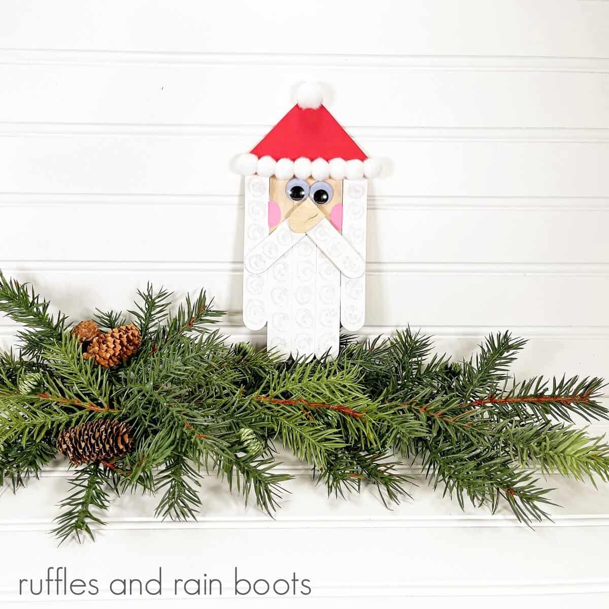 A square image of a completed Craft Stick Santa DIY on top of faux holiday greenery and pinecones against a white beadboard wood background.