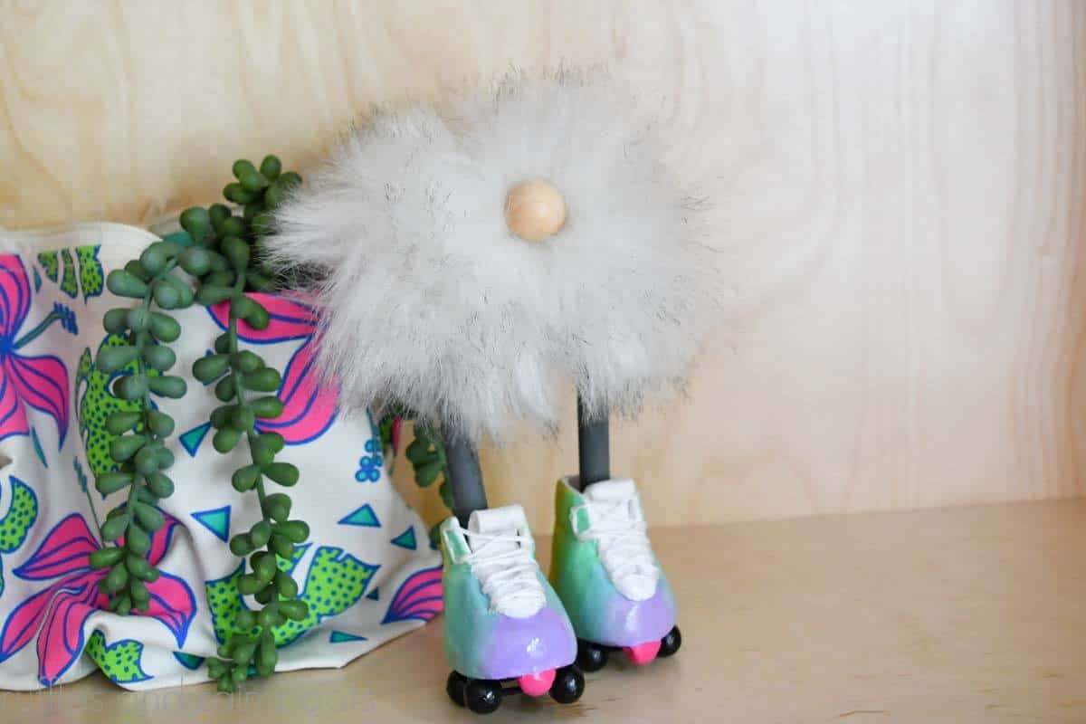 Horizontal image of a white pompom gnome with gray legs and rainbow roller skates next to a tropical bag and succulent.