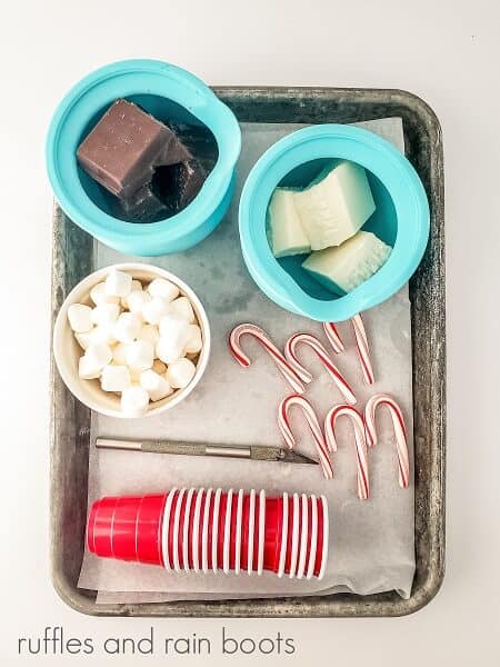 A small silicone cup with dark and another filled with white chocolate, a small ramekin filled with mini marshmallows, mini candy canes, small red plastic shot glasses and an xacto knife on parchment paper on a metal baking tray against a white backdrop.