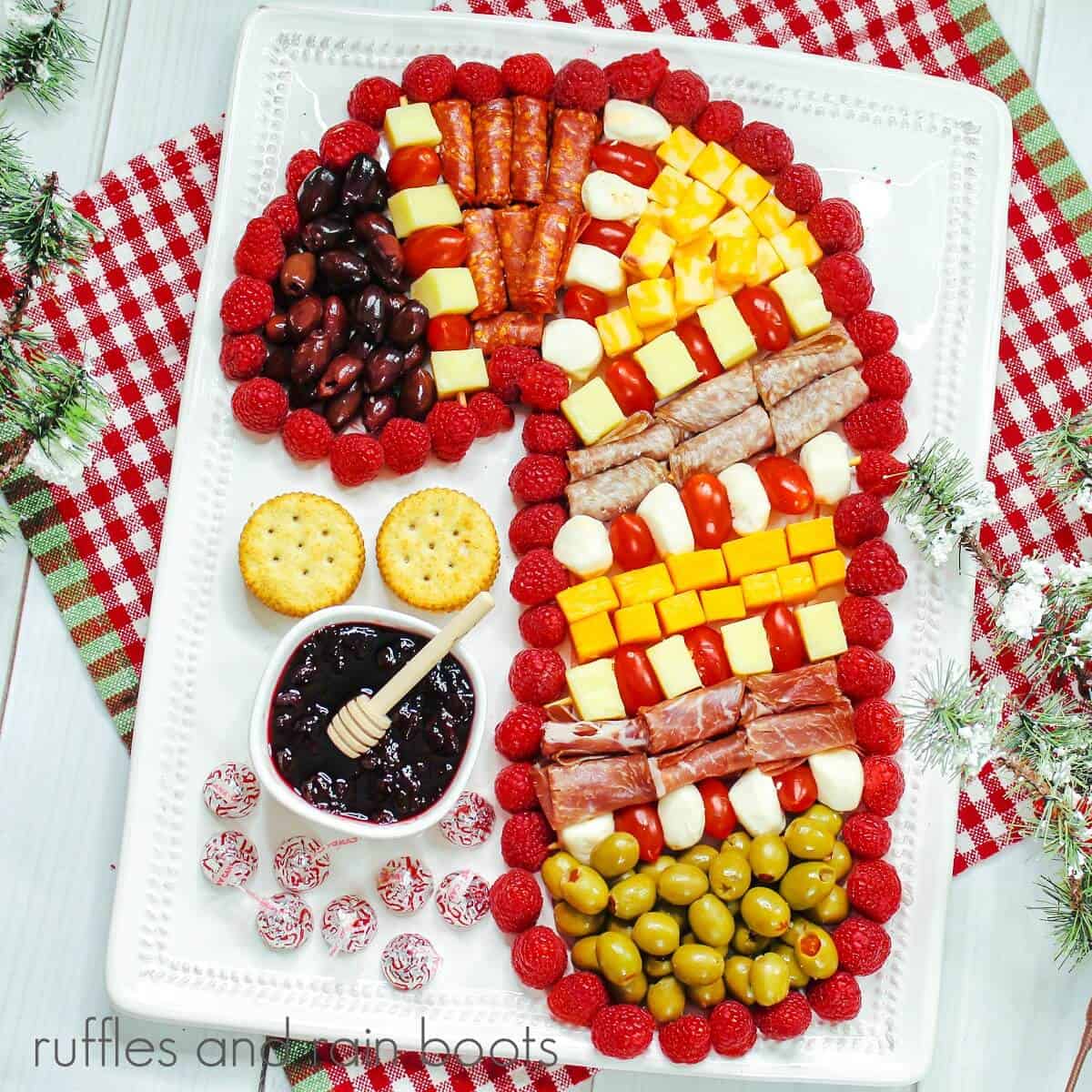 A square image of the meat, cheese, olives and fruit charcuterie board in the shape of a candy cane, on a white plate against a red and white gingham towel on a weathered white wood backdrop, next to faux winter greenery.