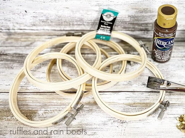 Several wooden embroidery hoops next to a bottle of brown paint and a paint brush against a white weathered wood background.