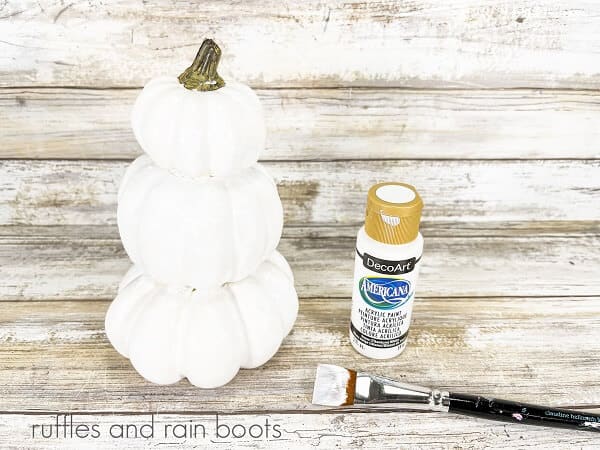 Stacked pumpkin snowman craft next to a bottle of white acrylic paint and a paintbrush, against a white weathered wood backdrop.