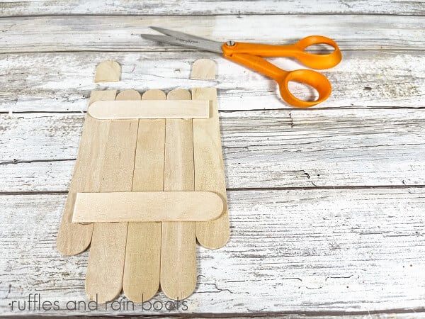 5 jumbo craft sticks, glued together, next to a pair of orange-handles scissors against a white weathered wood background.