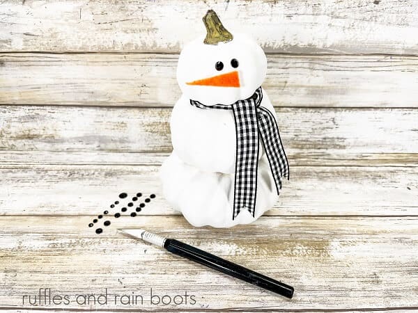 Stacked pumpkin snowman craft next to a xacto knife and self adhesive rhinestones, against a white weathered wood backdrop.