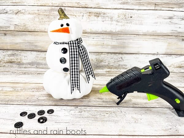 Stacked pumpkin snowman craft next to a hot glue gun and 6 black buttons, against a white weathered wood backdrop.