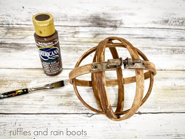 A craft store embroidery hoop orb next to a bottle of brown paint and a paintbrush on a white weathered wood background.