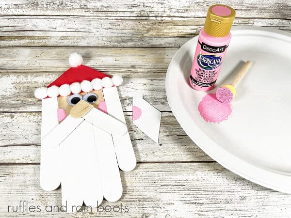 A Craft Stick Santa DIY next to a bottle of pink acrylic pain and a foam paint dauber on a foam plate against a white weathered wood background.