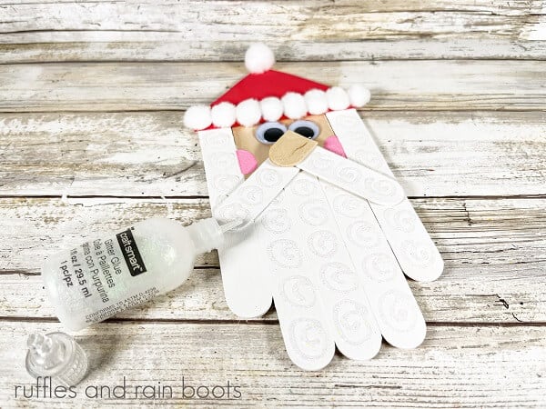 A completed Craft Stick Santa DIY next to a bottle of glitter glue against a white weathered wood background.
