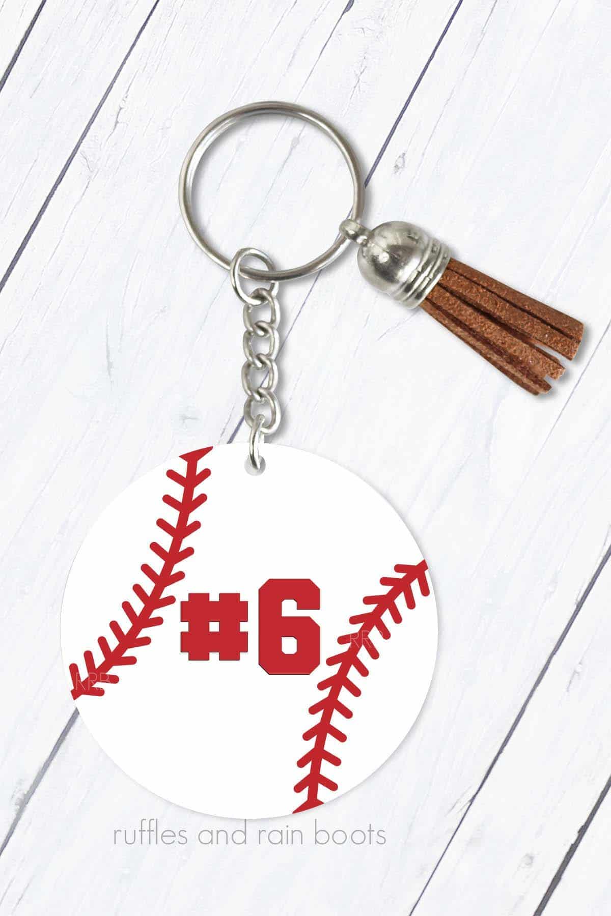 Vertical image of a baseball keychain with a brown tassel on a white wood background.