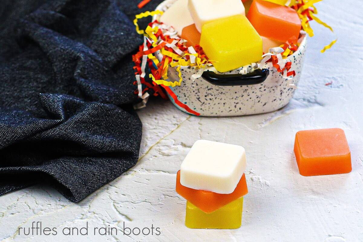A small black and white container filled with orange, white and yellow sugar scrub cubes and candy corn shredded paper next to more sugar scrub cubes and a black towel against a white marble background.