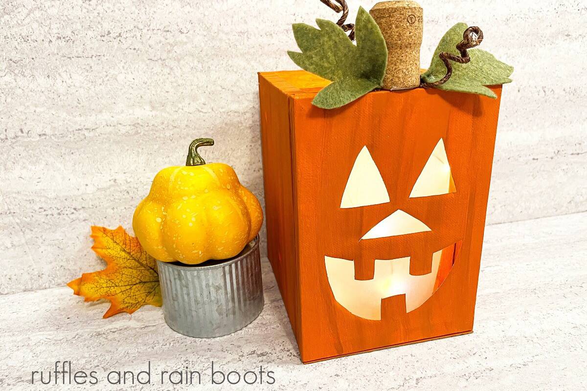 A horizontal Completed lighted canvas Jack-O-Lantern craft with a fall squash on a metal tin next to a fall leaf on the left against a white and grey background.