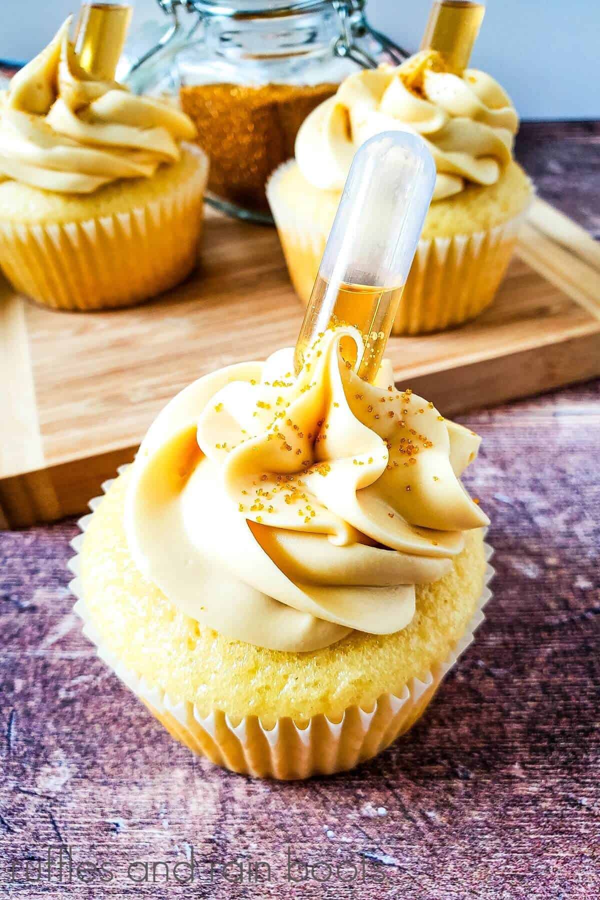 A cognac cupcake in front of a wooden cutting board with 2 additional cupcakes on it, against a weathered wood background.