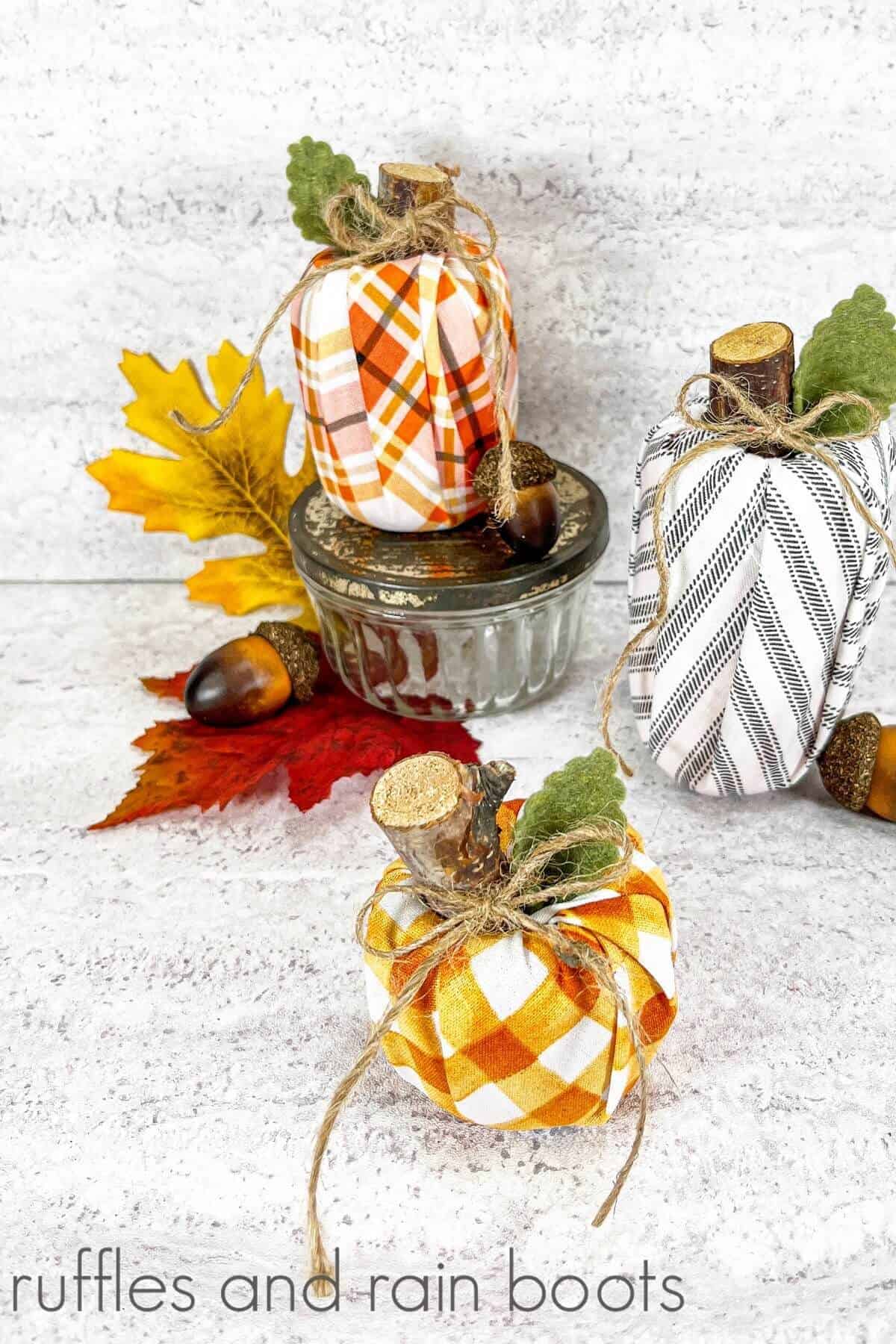 A vertical image of three wrapped Dollar Store pool noodle pumpkins next to faux fall leaves acorns and a small glass jar with a galvanized metal lid on a white and grey background.