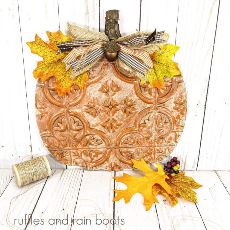A square image of a completed Dollar Tree Faux Tin Pumpkin next to a spool of off white thread, silk fall leaves and fall berries against a white wood background