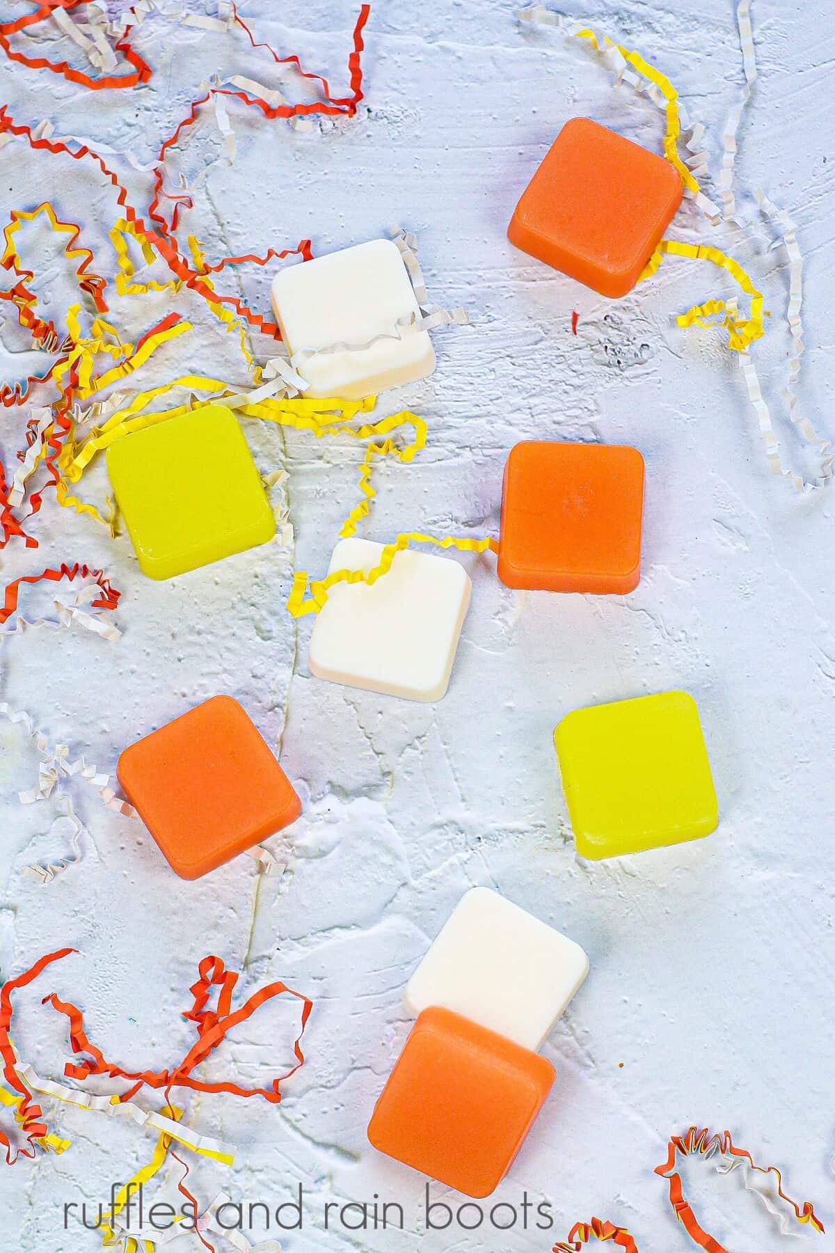 Several Candy Corn Sugar Scrub Cubes next to candy corn colored shredded paper against a white concrete background.