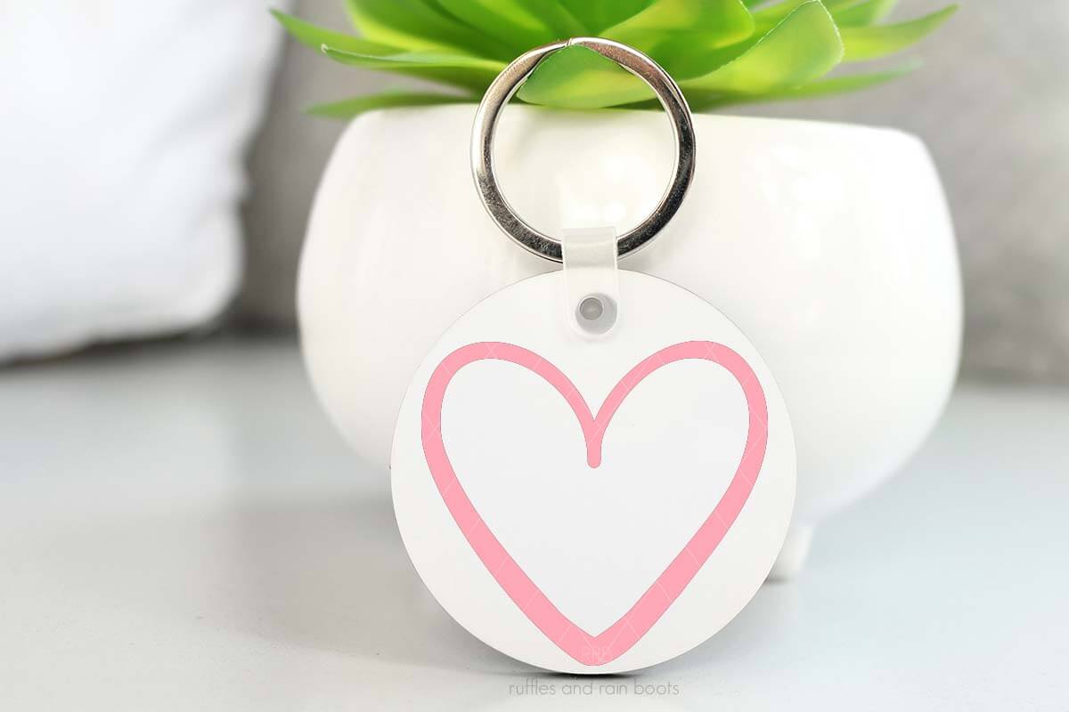 Horizontal image of white keychain with pink heart SVG propped against a small plant.