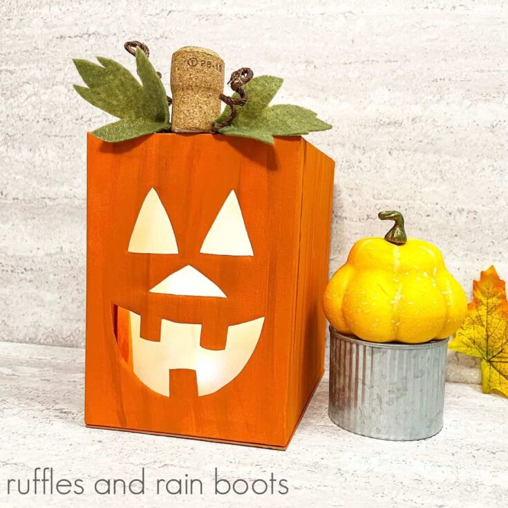 A square image of a lighted canvas Jack-O-Lantern next to a fall squash on a metal tin next to a fall leaf against a white and grey background.