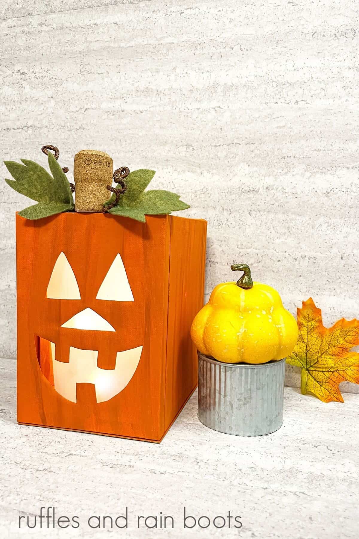 Completed lighted canvas Jack-O-Lantern craft next to a fall squash on a metal tin next to a fall leaf against a white and grey background.