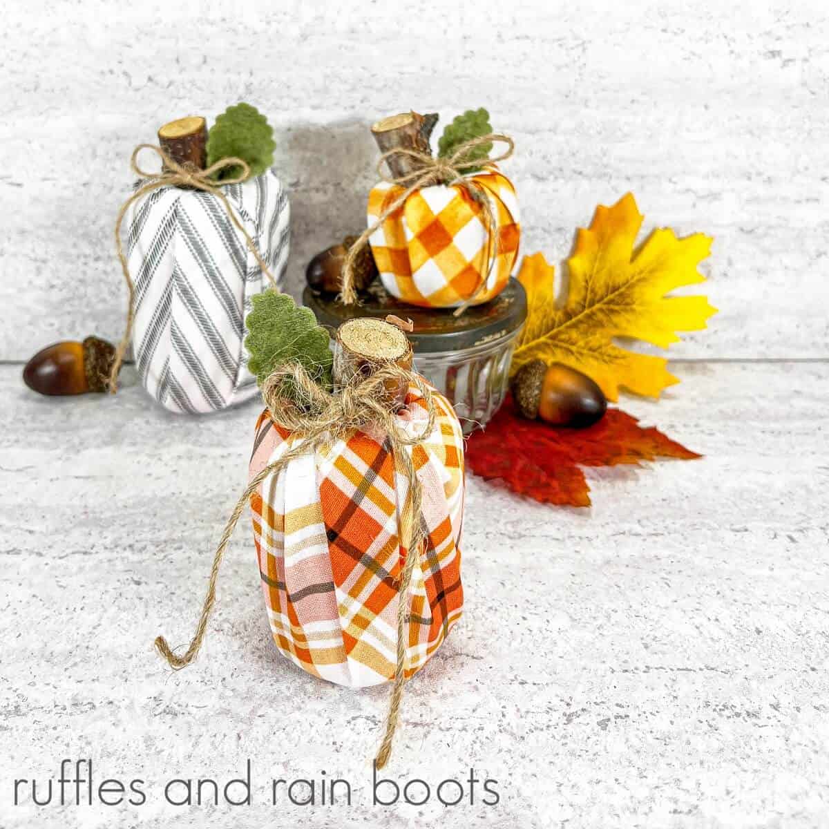 Three wrapped Dollar Store pool noodle pumpkins, one in black and white, one orange and white and one orange plaid, next to faux fall leaves acorns and a small glass jar with a galvanized metal lid on a white and grey background.