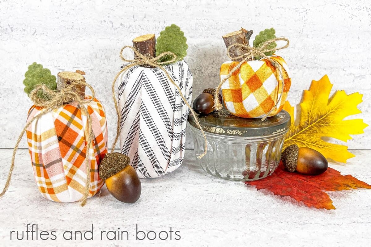 Three wrapped Dollar Store pool noodle pumpkins next to faux fall leaves acorns and a small glass jar with a galvanized metal lid on a white and grey background.