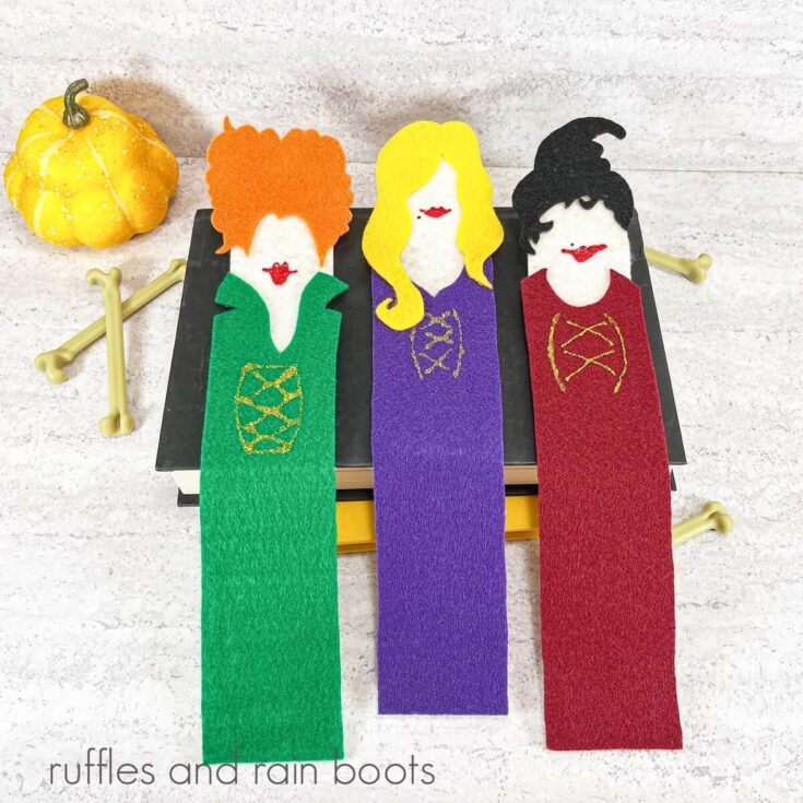 Vertical image of three Sanderson Sisters bookmarks on a stack of books, next to 4 faux bones and a yellow gourd against a white and grey background.