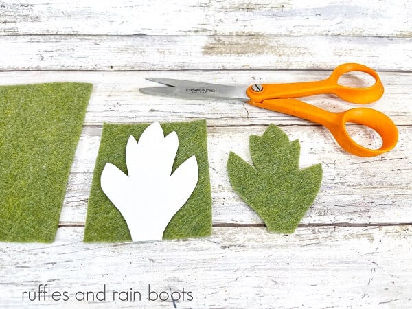 A piece of green felt with a leaf template on top, next to another piece of felt and a pair of scissors against a white weathered wood background.