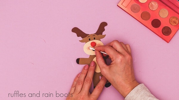 Crafter holding a q-tip, applying make-up to the reindeer's face, next to an eyeshadow palette against a purple background.