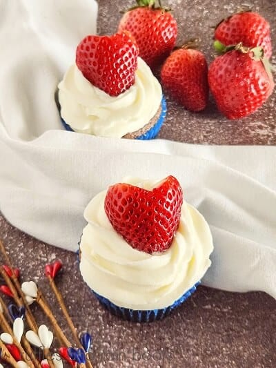 Two frosted blueberry cupcakes with strawberries on top, next to a bunch of fresh strawberries, next to holiday decorations against a brown background.