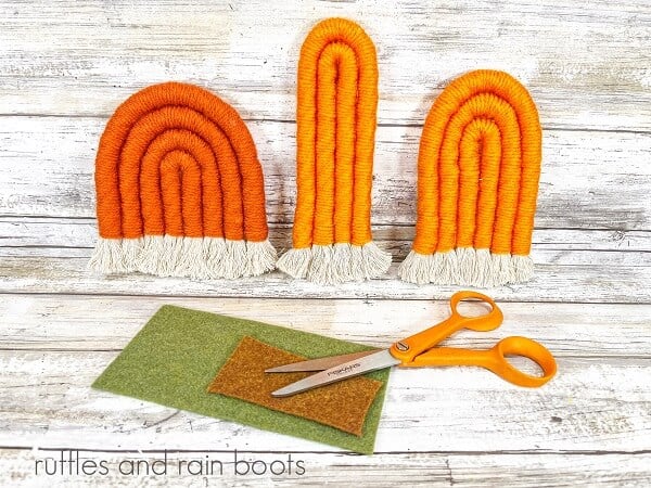 Three rope pumpkins next to green and brown felt and a pair of scissors against a white weathered wood background.