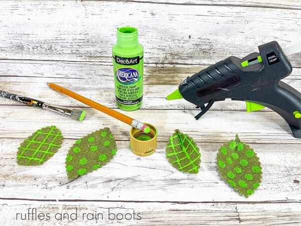 Four painted leaves next to a bottle of green paint, a hot glue gun, pencil and a paintbrush against a white weathered wood background.