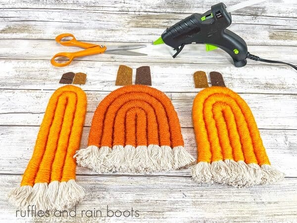 Three rope pumpkins next to brown felt for the stems next to a hot glue gun and scissors against a white weathered wood background.