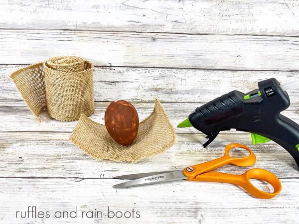 A painted Easter egg on a rectangle of burlap ribbon next to a roll of burlap ribbon, a pair of scissors and a hot glue gun against a white weathered wood background.