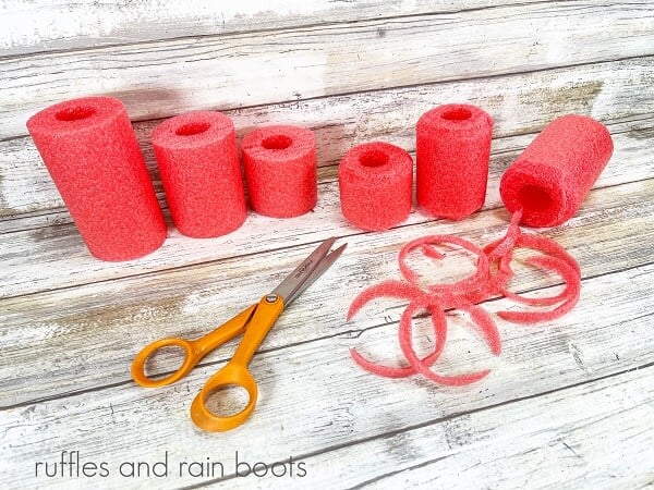 Six pool noodles next to a pair of scissors against a white weathered wood background.