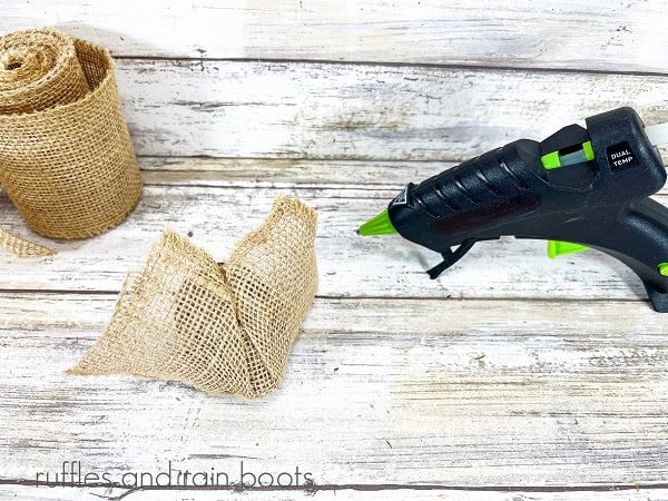 A Easter egg being wrapped in burlap ribbon, next to a hot glue gun against a white weathered wood background.