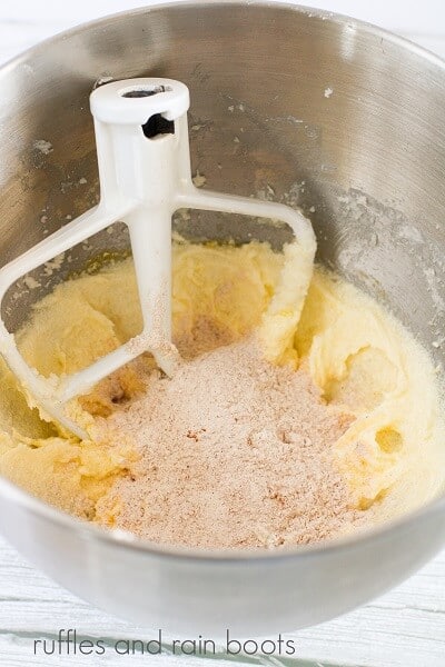 A large metal mixing bowl with a paddle attachment inside, on top of the wet and dry snickerdoodle cupcake ingredients.