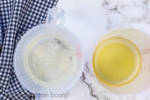 Vertical image of a measuring cup filled with melted clear soap next to a measuring cup with Shea butter, next to a black and white checkered towel against a white marble background.