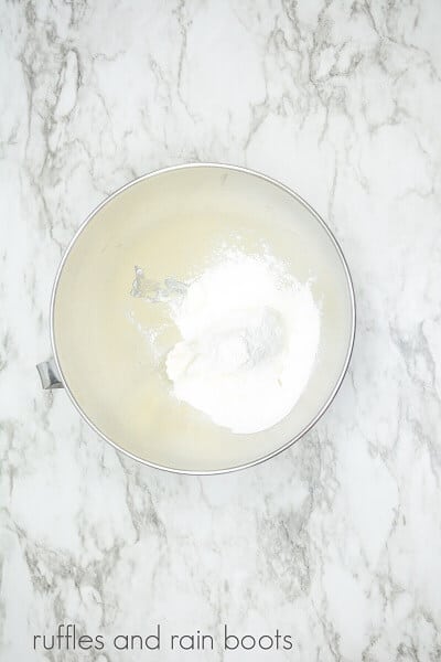 Vertical image of a stainless steel mixing bowl with dry cupcake ingredients against a white marble background.