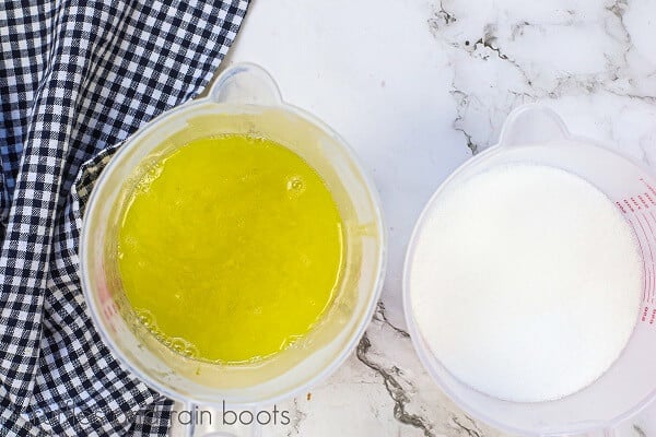 Yellow melted soap in a measuring cup next to a measuring cup with sugar next to a black and white checkered towel on a white marble background.