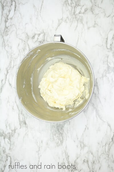 A stainless steel mixing bowl of banana buttercream frosting on a white marble background.