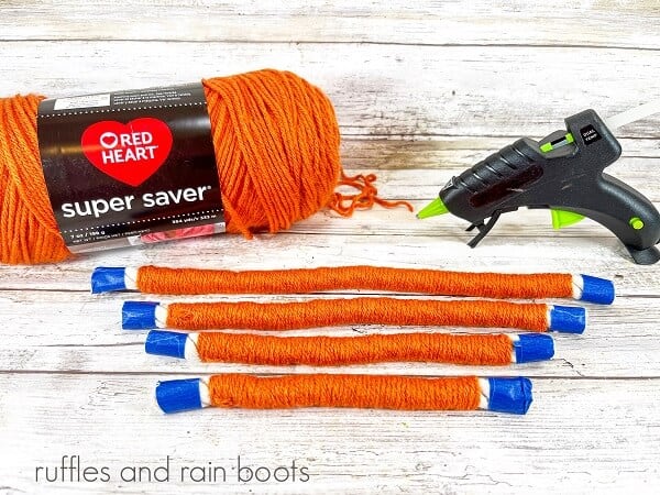 Yarn wrapped pieces of rope with painter's next to a hot glue gun and a skein of orange yarn against a white weathered wood background.
